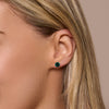Close-up of a woman's ear wearing a green gemstone stud earring on a beige background